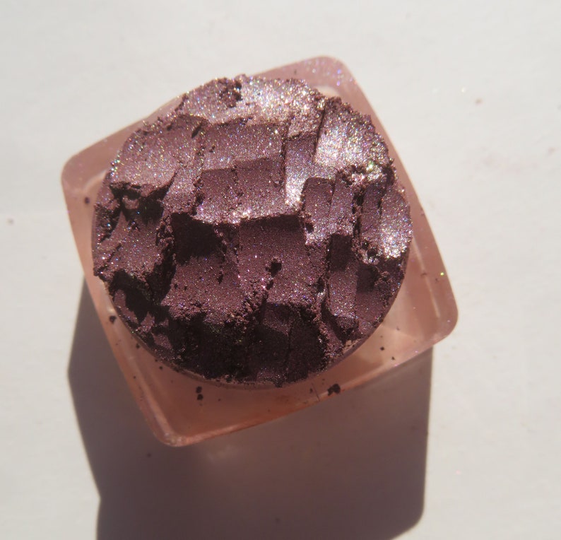 May include: A close-up of a small, round container of purple eyeshadow with a shimmery finish. The eyeshadow is in a clear container with a pink lid.