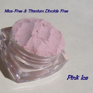 May include: A small, clear, square-shaped cosmetic jar filled with light pink powder. The text "Mica-Free & Titanium Dioxide Free" appears at the top, and the text "Pink Ice" is printed in the bottom right corner of the image.