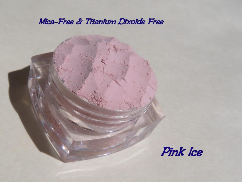 May include: A small, clear, square-shaped cosmetic jar filled with light pink powder. The text "Mica-Free & Titanium Dioxide Free" appears at the top, and the text "Pink Ice" is printed in the bottom right corner of the image.