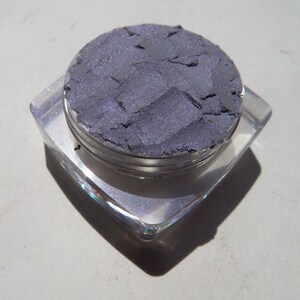May include: A small, round container of dark purple eyeshadow with a silver lid. The eyeshadow is in a pressed powder form and has a shimmery finish.
