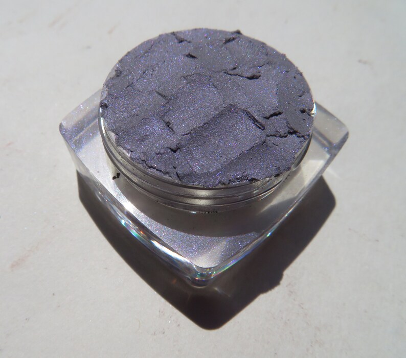 May include: A small, round container of dark purple eyeshadow with a silver lid. The eyeshadow is in a pressed powder form and has a shimmery finish.