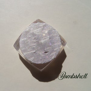 Op de afbeelding: Een kleine, heldere, vierkante container met een glinsterend, licht zilverkleurig cosmetisch product. Het product heeft een textuur en glinstert in het licht. Het woord "Bombshell" staat in cursief schrift rechtsonder.