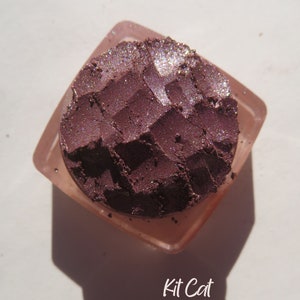 May include: A close-up of a small, round container of purple eyeshadow with a shimmery finish. The container is made of clear plastic and has a textured surface. The eyeshadow is labeled "Kit Cat".