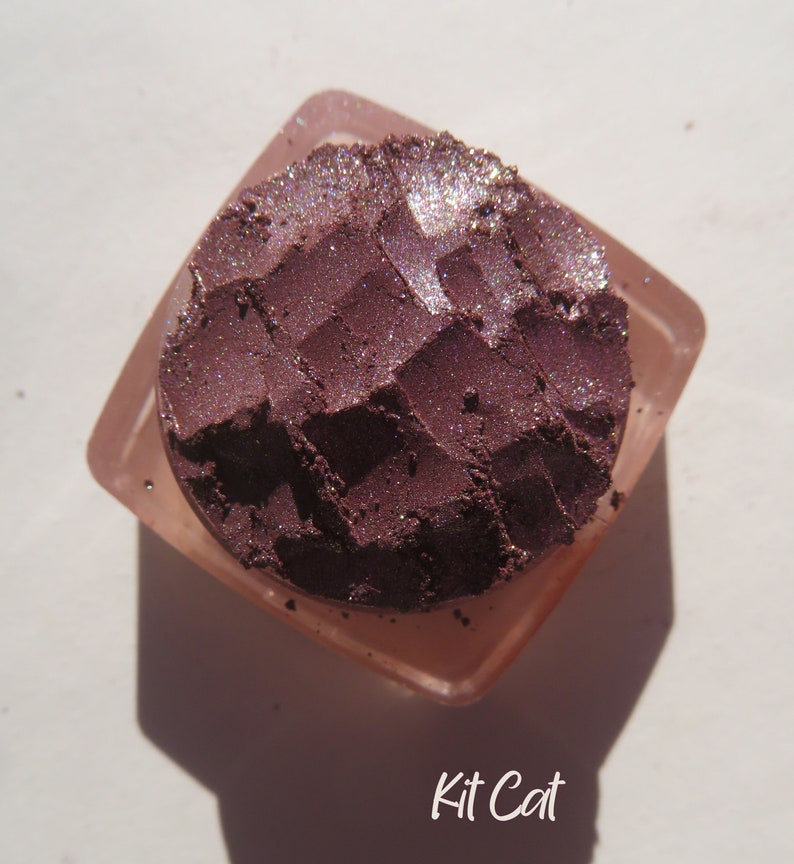 May include: A close-up of a small, round container of purple eyeshadow with a shimmery finish. The container is made of clear plastic and has a textured surface. The eyeshadow is labeled "Kit Cat".