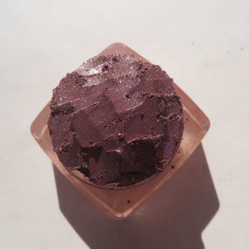 May include: A small container of loose purple eyeshadow with a shimmery finish. The eyeshadow is in a clear square container with a pink lid.