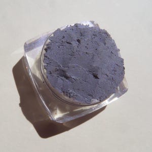 May include: A small, clear plastic container filled with a dark gray eyeshadow powder.