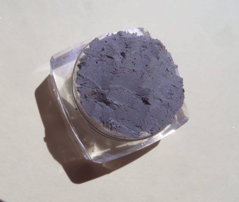 May include: A small, clear plastic container filled with a dark gray eyeshadow powder.