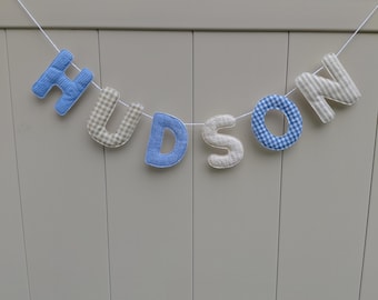 May include: A fabric banner spelling the name HUDSON, featuring individual stuffed letters strung together on a white cord. The letters alternate between a blue and white gingham pattern and a beige and white gingham pattern.