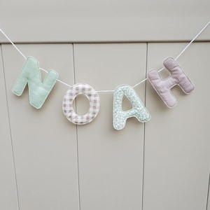 Puede incluir: Un cartel con el nombre "NOAH" hecho de letras de tela. Las letras tienen diferentes patrones y colores, incluyendo verde menta, cuadros, floral y rayas. El cartel est&aacute; colgado de una cuerda blanca sobre un fondo neutro.