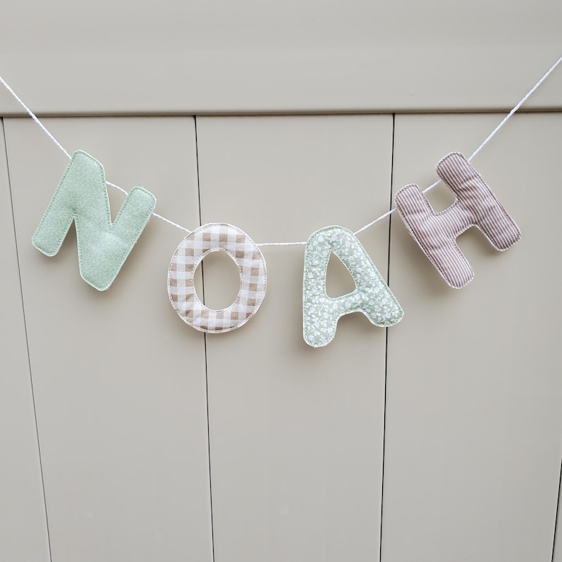 Puede incluir: Un cartel con el nombre "NOAH" hecho de letras de tela. Las letras tienen diferentes patrones y colores, incluyendo verde menta, cuadros, floral y rayas. El cartel est&aacute; colgado de una cuerda blanca sobre un fondo neutro.
