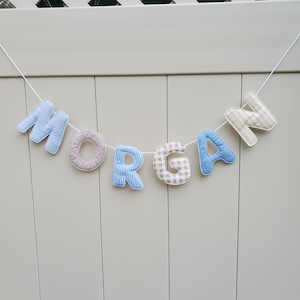 Puede incluir: Un cartel con la palabra "MORGAN" con letras de tela en varios patrones y colores. Las letras son de cuadros azules, rayas marrones y blancas, rayas azules, cuadros beige y blancos y azules. El cartel est&aacute; ensartado en una cuerda blanca.