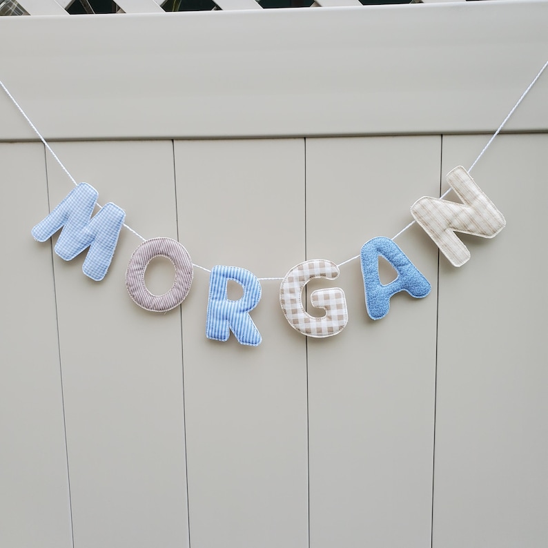 Puede incluir: Un cartel con la palabra "MORGAN" con letras de tela en varios patrones y colores. Las letras son de cuadros azules, rayas marrones y blancas, rayas azules, cuadros beige y blancos y azules. El cartel est&aacute; ensartado en una cuerda blanca.