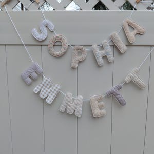Puede incluir: Un bander&iacute;n de tela hecho a mano con las letras "Sophia Emmett". Las letras, con diferentes patrones, est&aacute;n ensartadas en una cuerda blanca y cuelgan sobre un fondo gris claro, ideal para la decoraci&oacute;n de la habitaci&oacute;n de los ni&ntilde;os.