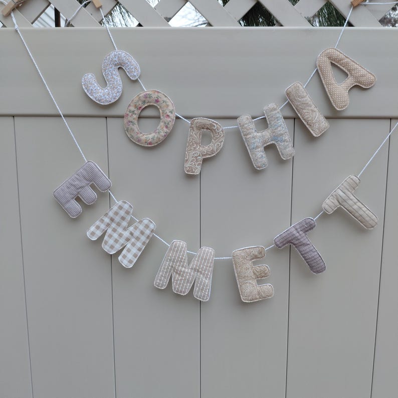 Puede incluir: Un bander&iacute;n de tela hecho a mano con las letras "Sophia Emmett". Las letras, con diferentes patrones, est&aacute;n ensartadas en una cuerda blanca y cuelgan sobre un fondo gris claro, ideal para la decoraci&oacute;n de la habitaci&oacute;n de los ni&ntilde;os.