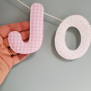 May include: Pink fabric banner with the letters "JO" in a gingham and polka dot pattern.