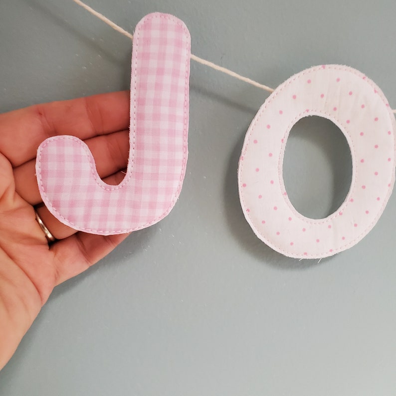 May include: Pink fabric banner with the letters "JO" in a gingham and polka dot pattern.