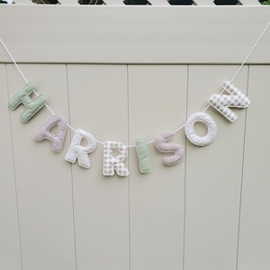Puede incluir: Una guirnalda de tela con el nombre "HARRISON" en varios colores pastel y patrones. Las letras est&aacute;n cosidas a una cuerda blanca, creando una guirnalda decorativa. El fondo es una valla de madera de color claro.