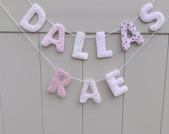 May include: A fabric letter banner spelling the name DALLAS RAE in two rows. The letters are crafted from various shades of pink and white fabric, featuring patterns including floral prints, gingham checks, and solid textures, strung on white cord.