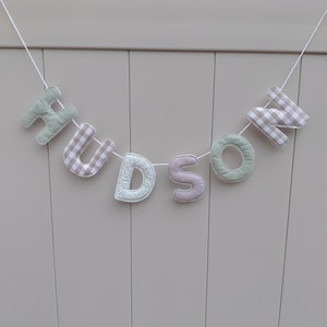 Puede incluir: Una guirnalda de tela con la palabra "HUDSON". Las letras son de varios colores y estampados, incluyendo verde, cuadros y tela a rayas, ensartadas en un cord&oacute;n blanco. La pancarta se muestra sobre un fondo neutro de color claro.