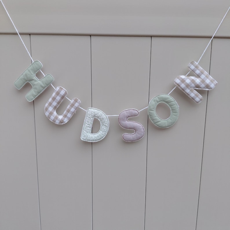 Puede incluir: Una guirnalda de tela con la palabra "HUDSON". Las letras son de varios colores y estampados, incluyendo verde, cuadros y tela a rayas, ensartadas en un cord&oacute;n blanco. La pancarta se muestra sobre un fondo neutro de color claro.