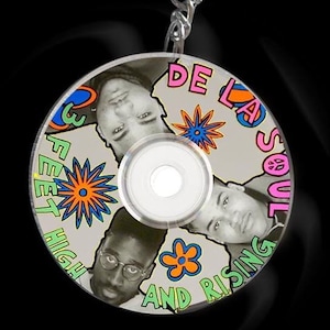 Op de afbeelding: Een zilveren sleutelhanger met een ronde, grijze schijf met een kleurrijk ontwerp. De schijf heeft de tekst "DE LA SOUL" en "3 FEET HIGH AND RISING" in een roze, groene en oranje lettertype. Er zijn drie gezichten van mensen op de schijf.