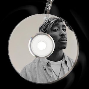 Op de afbeelding: Een zilveren sleutelhanger met een ronde metalen ring en een ketting die aan een cd is bevestigd. De cd heeft een zwart-witfoto van een persoon die een bandana en een shirt draagt. De persoon kijkt naar de camera.