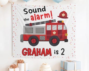 Puede incluir: Una pancarta blanca con una ilustración de camión de bomberos rojo y el texto "Sound the alarm!" y "GRAHAM is 2". La pancarta está decorada con confeti y globos. Una tarta y regalos están en primer plano.