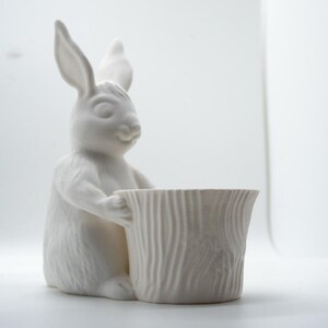Op de afbeelding: Witte keramische konijnenfiguur die een witte keramische plantenpot met houtnerf vasthoudt.