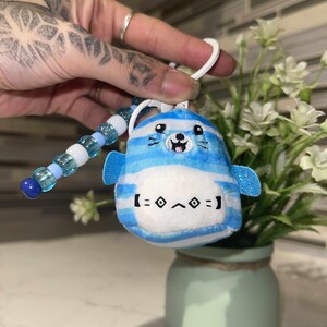 May include: A blue and white plush seal keychain with a smiling face and small blue flippers. The keychain has a white plastic loop and a beaded blue and white accent. The seal has the text "[= ◊ ^ ◊ =]" on its belly.