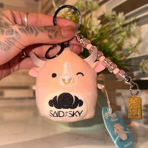 May include: A pink cow plush keychain with a black keyring and a beaded charm. The cow has small horns, a gold nose ring, and the text "SAID SKY" on its belly. A tag with the name "Howland" is attached.