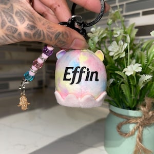 May include: A colorful keychain with a plush, round charm. The charm is tie-dye with the word "Effin" in black. The keychain has a beaded strand with a gold-colored Hamsa hand charm. The background is blurred.