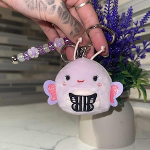 May include: A plush, lavender-colored keychain featuring a smiling creature with pink ears and a black and white butterfly design on its belly. The keychain includes a beaded charm with purple and white beads and a silver bell.