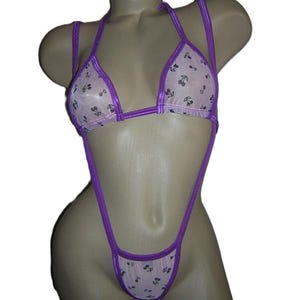 May include: A two-piece swimsuit in a light pink fabric with a cherry pattern. The edges and straps are a vibrant purple. The swimsuit is displayed on a mannequin.