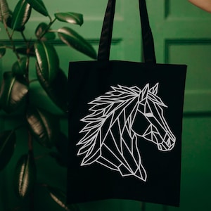 May include: Black tote bag featuring a white geometric horse head design. The tote bag has black handles and is held up against a green background. The horse head is a modern, minimalist illustration.