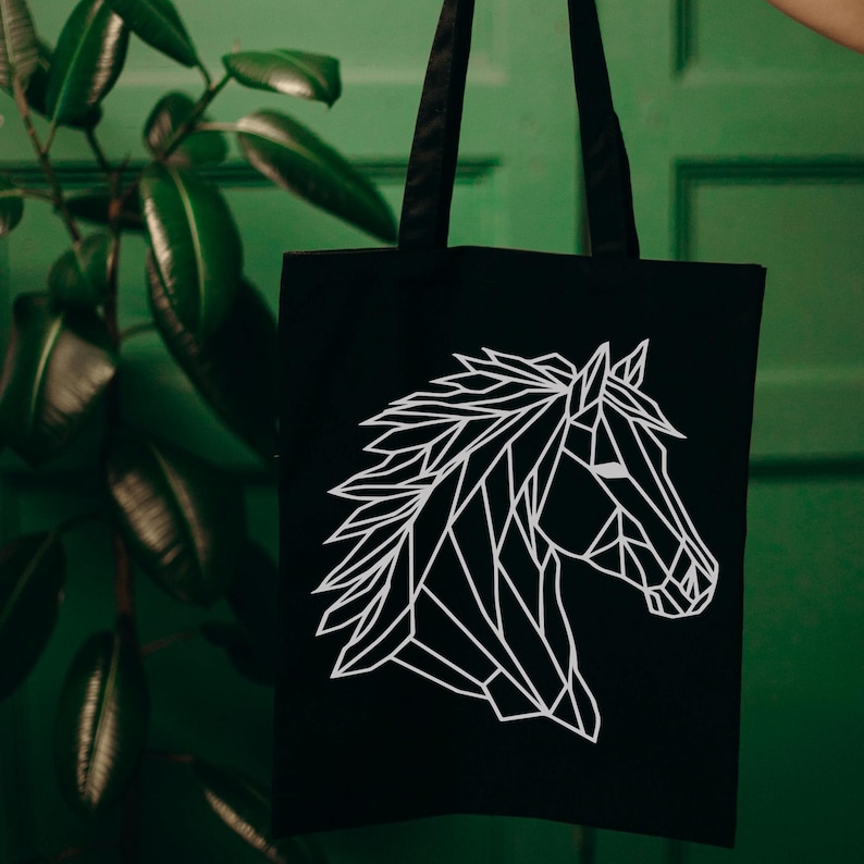 May include: Black tote bag featuring a white geometric horse head design. The tote bag has black handles and is held up against a green background. The horse head is a modern, minimalist illustration.