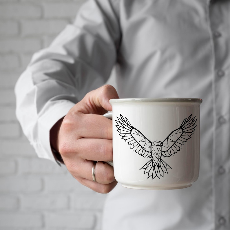 May include: White ceramic mug with a geometric black eagle design. The mug has a gray rim and handle. The eagle graphic features a polygonal design with outstretched wings. The mug is held by a person.