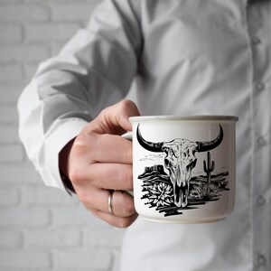 Może przedstawiać: Biały ceramiczny kubek z czarno-białą ilustracją czaszki byka, krajobrazu pustynnego i kaktusa. Kubek ma szarą obw&oacute;dkę i uchwyt. Na palcu osoby trzymającej kubek widoczny jest pierścionek.