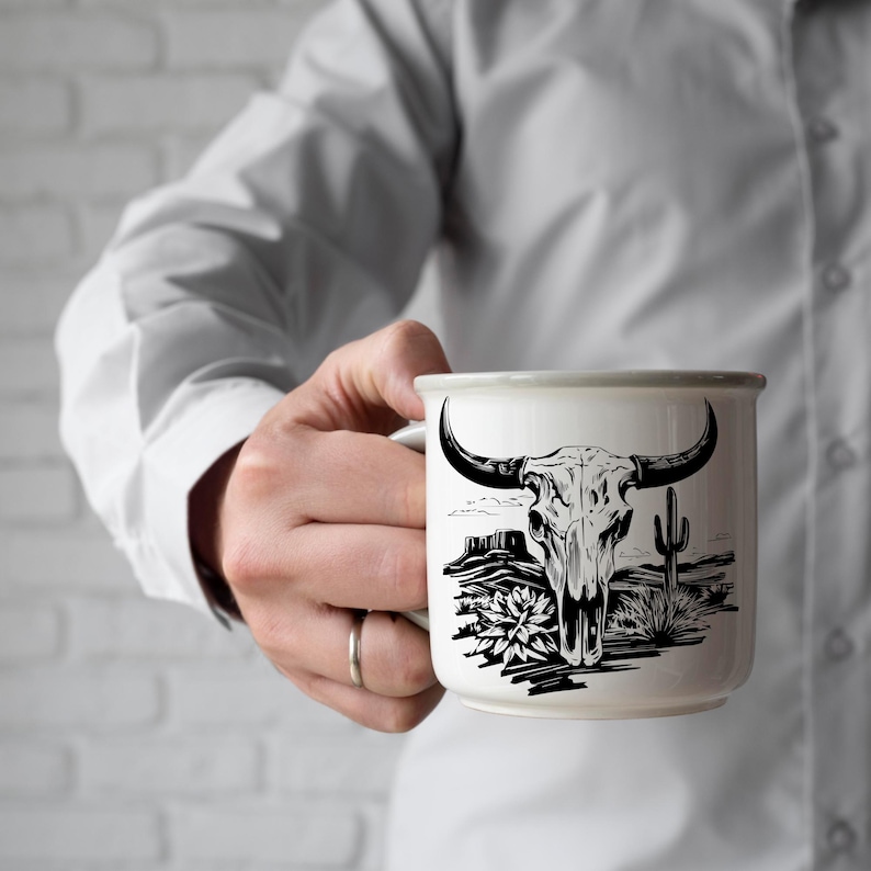 Może przedstawiać: Biały ceramiczny kubek z czarno-białą ilustracją czaszki byka, krajobrazu pustynnego i kaktusa. Kubek ma szarą obw&oacute;dkę i uchwyt. Na palcu osoby trzymającej kubek widoczny jest pierścionek.