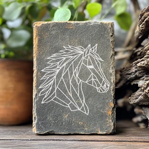 May include: A rectangular slate coaster featuring a geometric white outline of a horse's head. The coaster has a rough, textured surface with variations of gray and brown. The horse design is a modern, minimalist style.