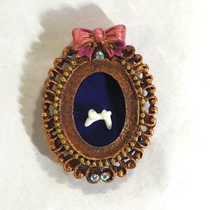 May include: A small, oval-shaped, gold-colored frame with a blue velvet background. A white tooth is displayed inside the frame. The frame is adorned with a pink bow with a rhinestone in the center.