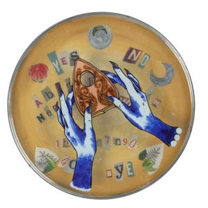 Puede incluir: Un plato decorativo redondo, de color dorado, con un diseño de tablero Ouija. El diseño incluye una plancheta, manos azules con uñas largas y negras, letras, números y las palabras "YES" y "NO".