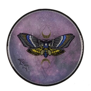 Puede incluir: Un diseño circular con borde negro, que presenta una ilustración de una polilla con una calavera. Las alas de la polilla son grises, azules y amarillas, sobre un fondo de galaxia púrpura. Las medias lunas doradas y las líneas geométricas completan el diseño.