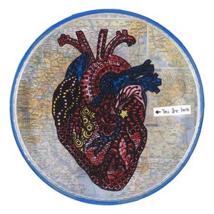 Puede incluir: Ilustración de un corazón colorido y estampado con diseños rojos, azules y negros, sobre un fondo de mapa. La obra de arte está enmarcada en un círculo azul, con el texto "You are here" y una flecha.