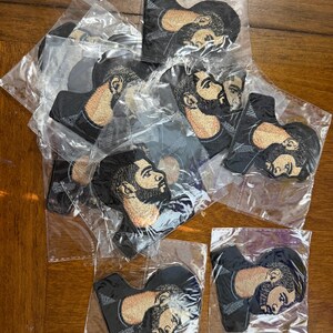 May include: Embroidered patches featuring a profile portrait of a man with a beard and stylish haircut. The patches are black with tan and brown details, packaged in clear plastic. The patches are likely for clothing or accessories.