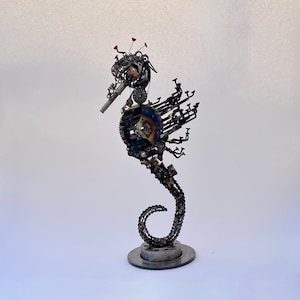 May include: A metal seahorse sculpture crafted from various machine parts. The seahorse features a curved body, a long snout, and a curled tail. The sculpture is primarily silver and gray, with hints of blue and red accents.