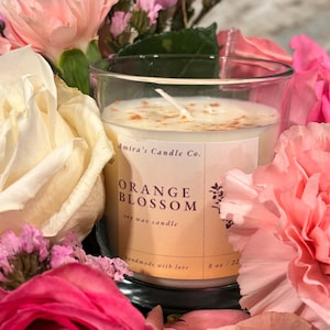 May include: A clear glass candle jar containing a white soy wax candle. The candle is labelled "Orange Blossom" and "Amira's Candle Co.". The jar is surrounded by pink and white flowers.