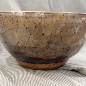 May include: A ceramic bowl with a rounded shape and a glossy finish. The upper portion features a mottled beige and tan glaze with dark brown speckles, transitioning into a darker brown band near the base, which reveals the raw, speckled clay.