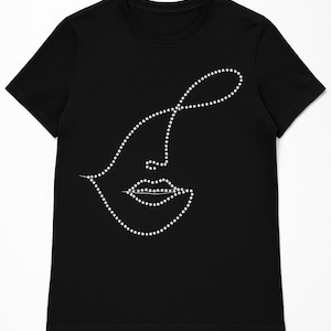 Puede incluir: Camiseta negra con un diseño minimalista de rostro. La cara está formada por pequeños puntos blancos, creando el contorno de una nariz, labios y un ojo abstracto. El diseño está centrado en la camiseta.