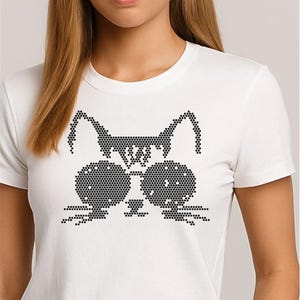 May include: White t-shirt featuring a black cat face design. The cat face is made of small black dots, with large round eyes and pointed ears. The shirt is a crew neck style.
