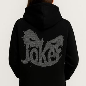 May include: Black hooded sweatshirt with the word "Joker" in a stylized design on the back. The word is composed of many small, light-colored dots. The sweatshirt has a hood and long sleeves.